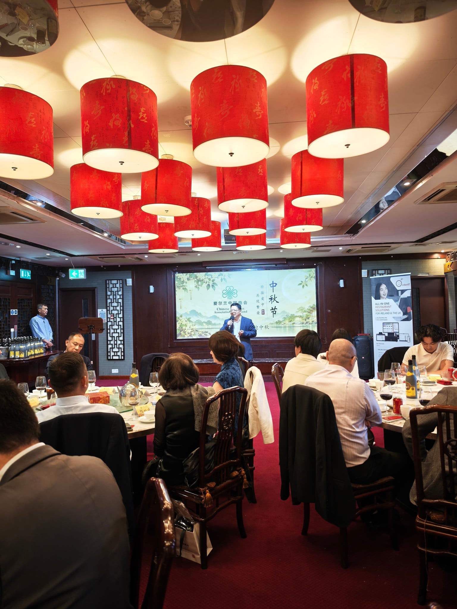 A speaker with a microphone in front of the Chamber's Mid-Autumn screen, beneath red lanterns, as guests listen from round tables.
