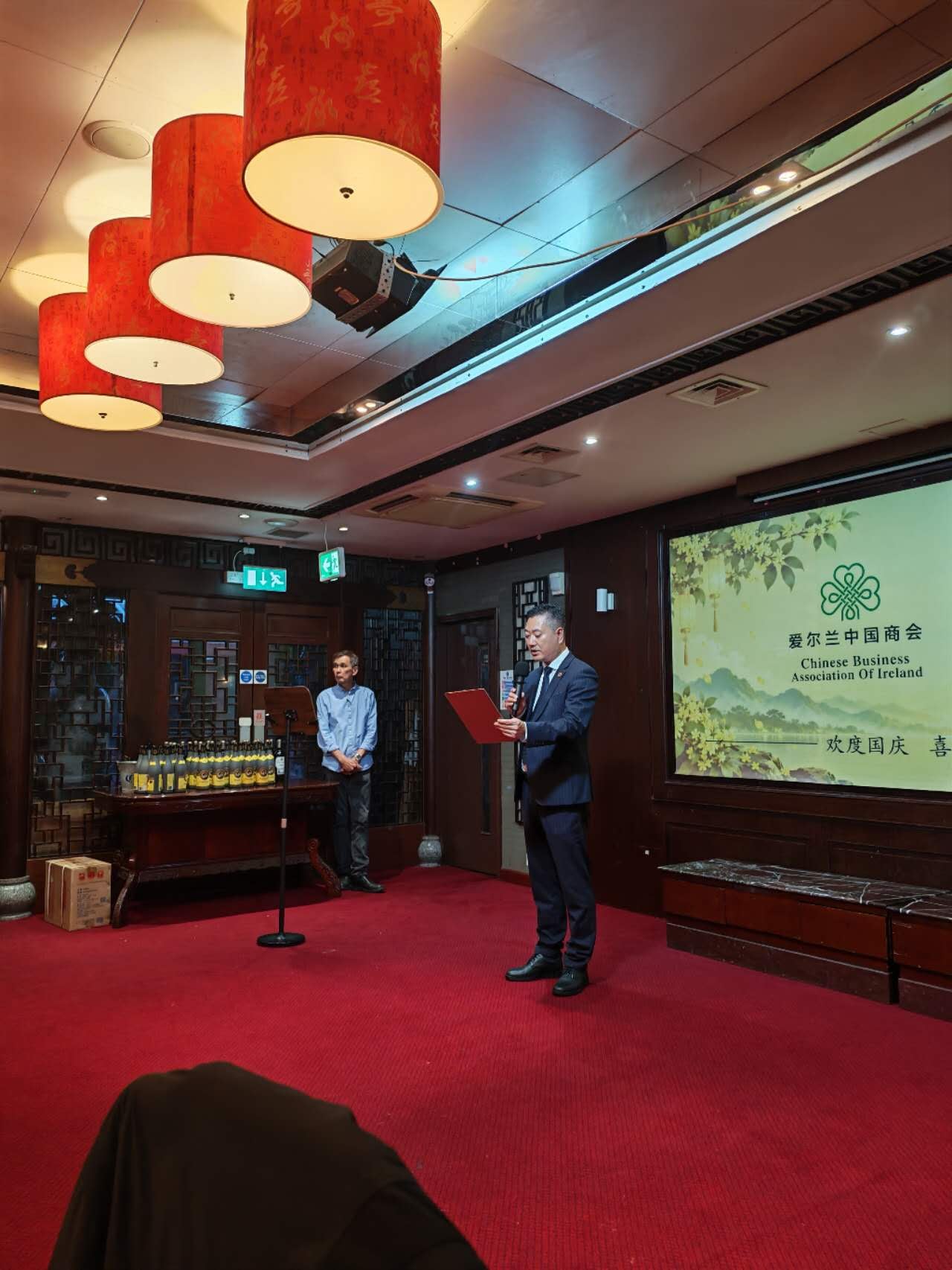 Yu Yongqiang, in a dark suit, reads from a red folder at a microphone, beside the Chamber's National Day and Mid-Autumn screen.