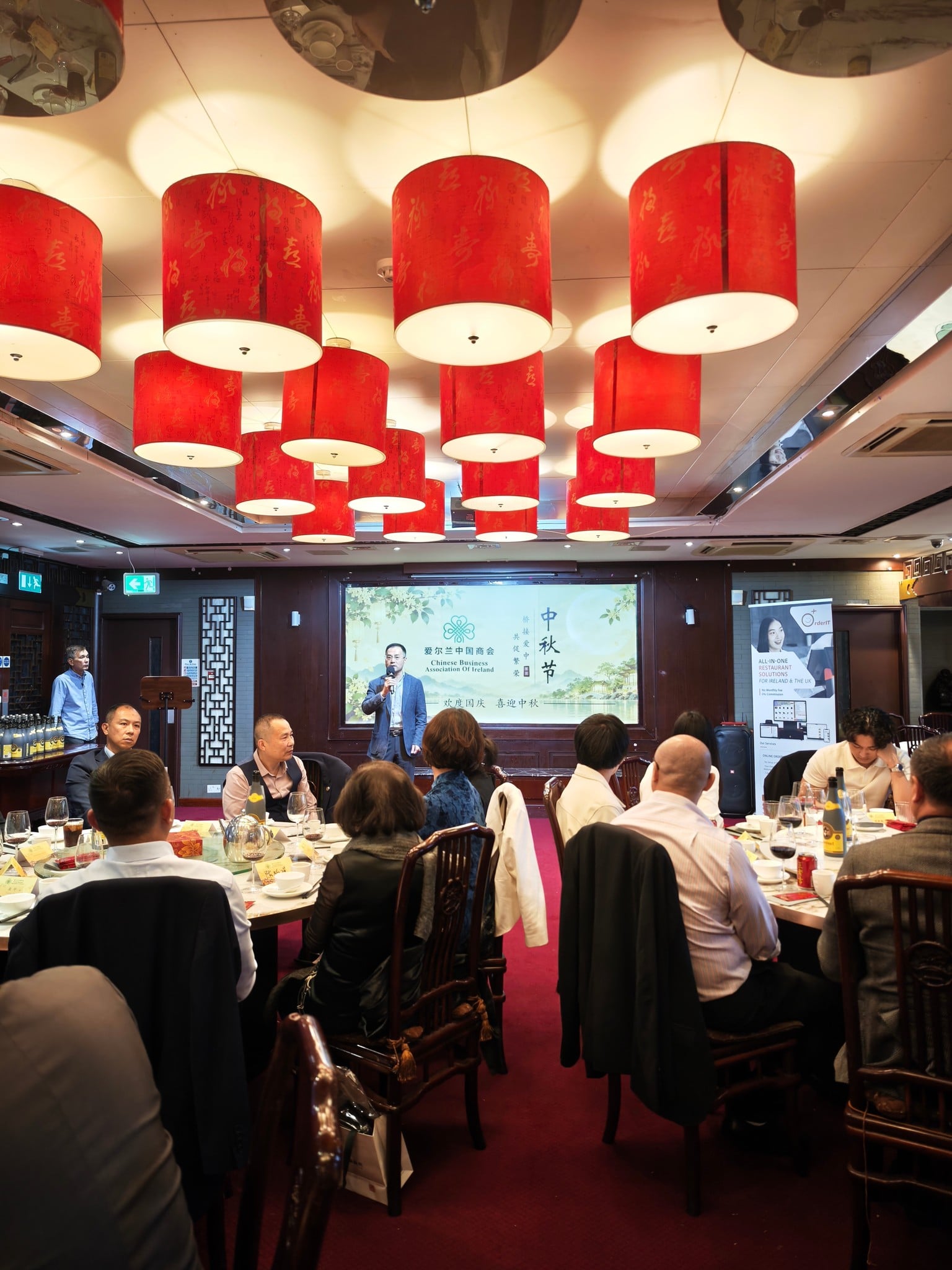 A second speaker addresses the room in front of the Chamber's “Celebrating National Day, Welcoming Mid-Autumn” screen, as guests listen from round tables.